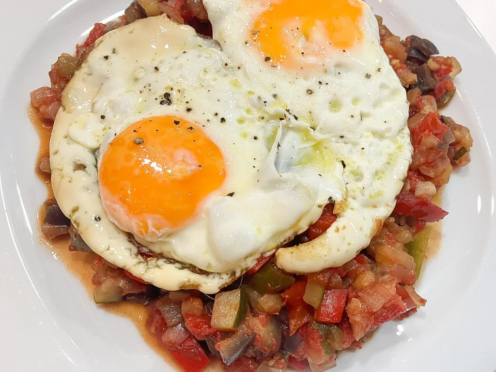 Pisto manchego con dos huevos fritos encima, servido en un plato blanco
